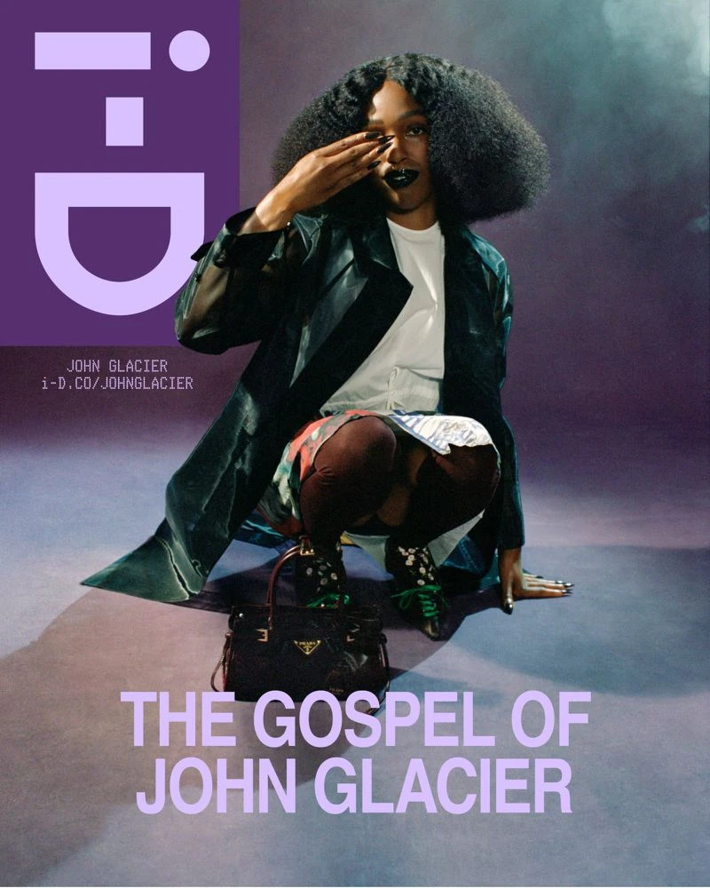 John Glacier crouches wearing a dark coat and white shirt with a black handbag in front of a purple backdrop