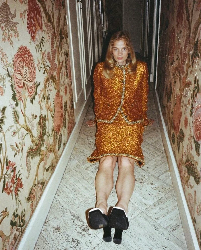 Model seated on a stone-tiled hallway floor between floral wallpaper walls wearing a gold textured skirt suit