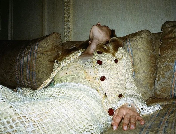 Model reclining on a patterned sofa wearing an open cream knit cardigan with dark buttons and a matching skirt