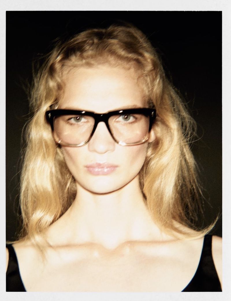 Close-up portrait of blonde woman wearing thick black-rimmed glasses against a dark background