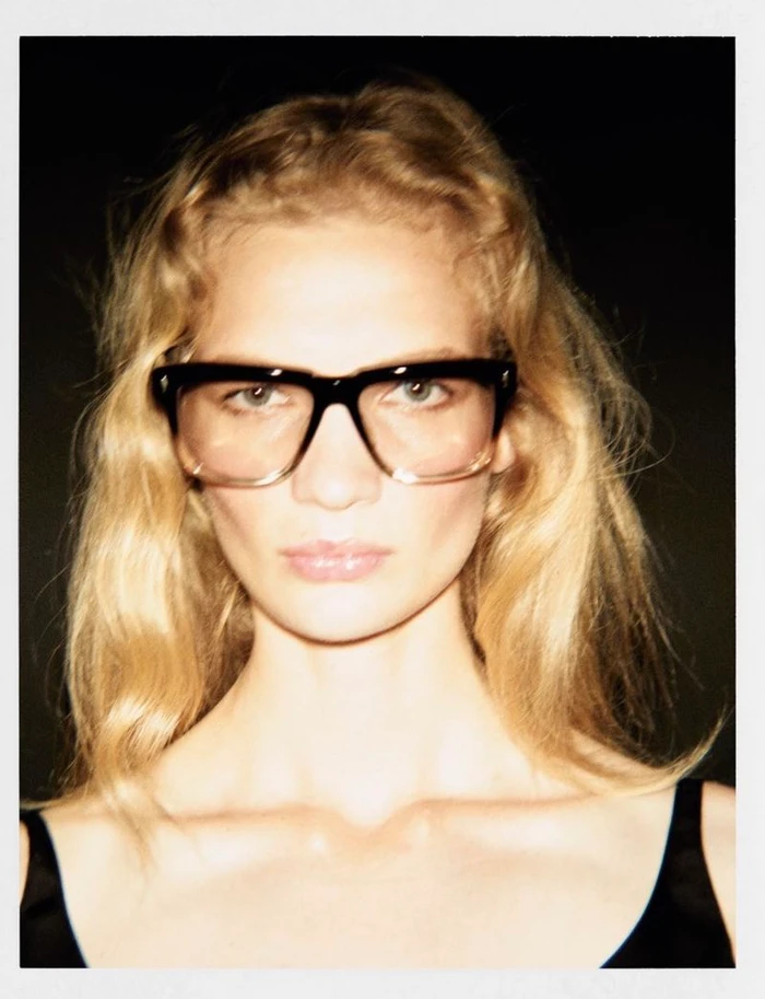 Close-up portrait of blonde woman wearing thick black-rimmed glasses against a dark background