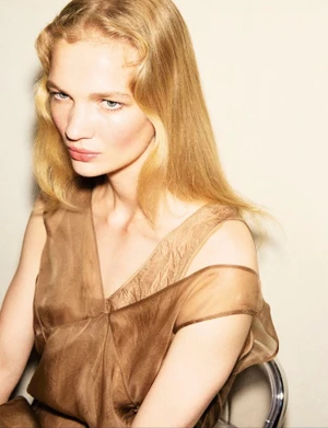 Model with long blonde hair wearing a sheer brown dress against a plain neutral background.