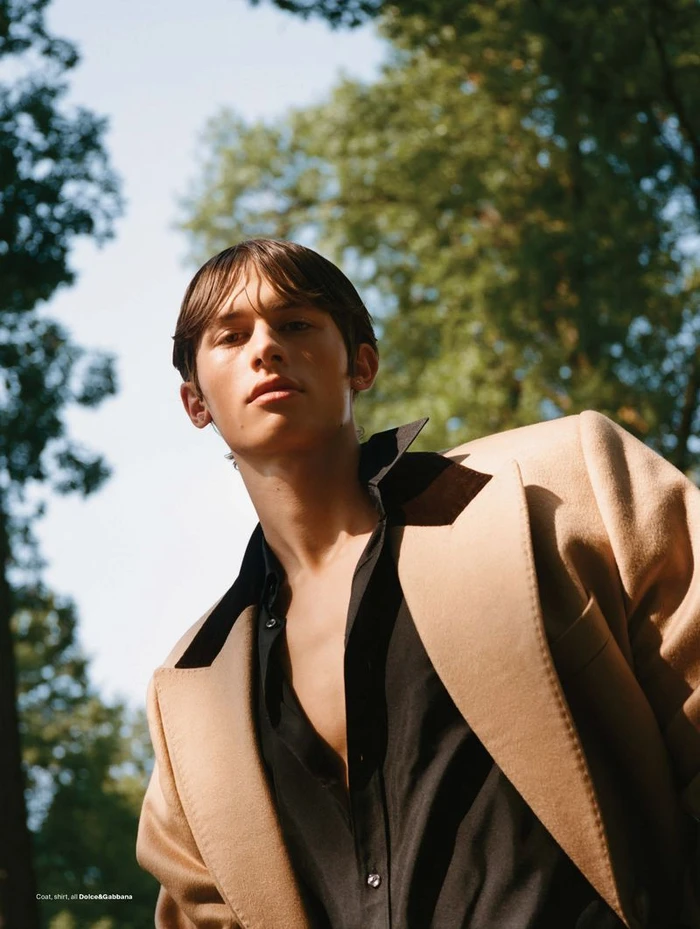 Model Josiah Turner looking up wearing a camel coat and black shirt outdoors against trees