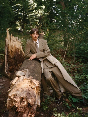 Model leaning against a large fallen log in a wooded forest wearing a tailored brown suit and tie.