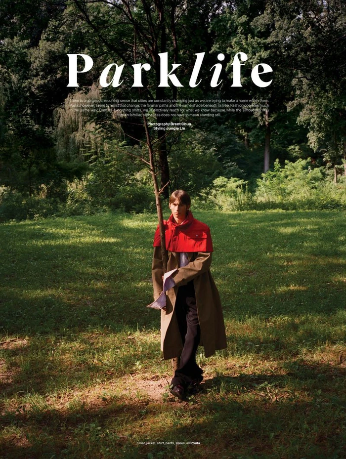 Model standing on a grassy path in a wooded park wearing a red and tan coat with dark trousers, photographed by Brent Chua.