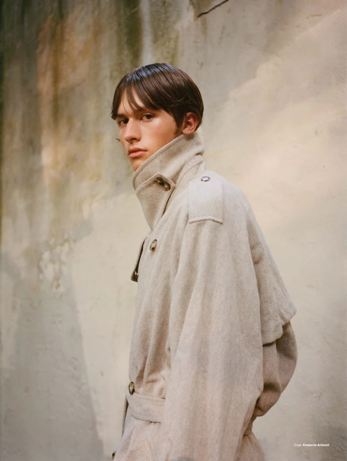 Model wearing a textured cream trench coat with an upturned collar outdoors against a mottled wall background.