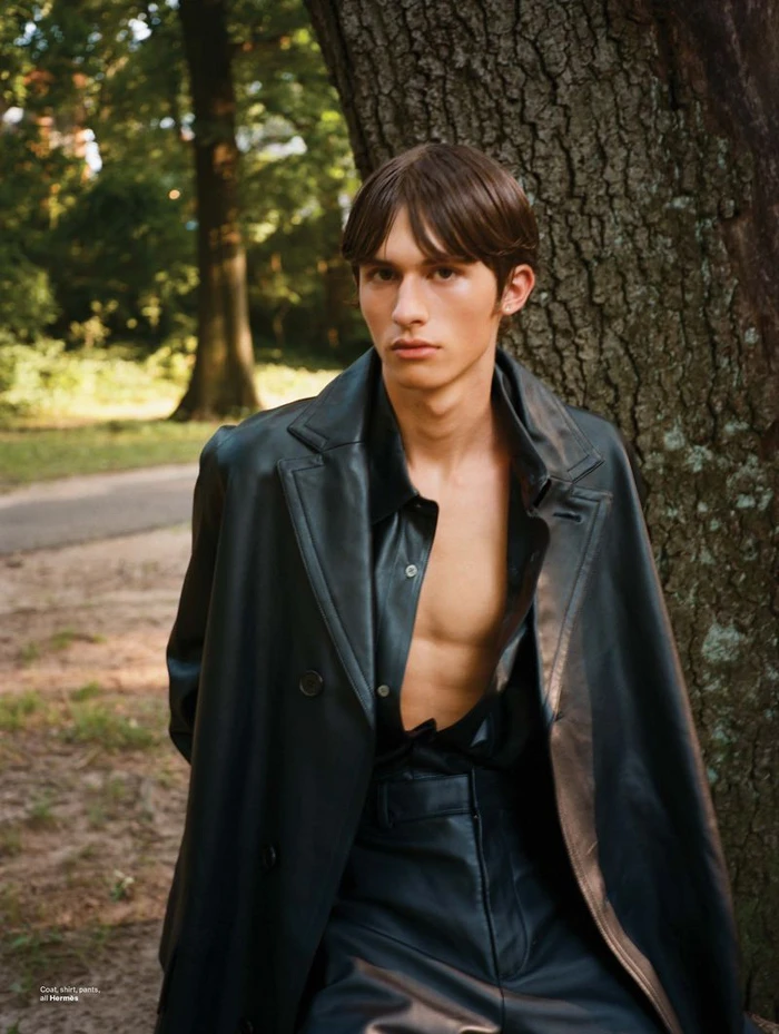 A young male model wearing a dark leather coat leaning against a large tree trunk in an outdoor park setting.