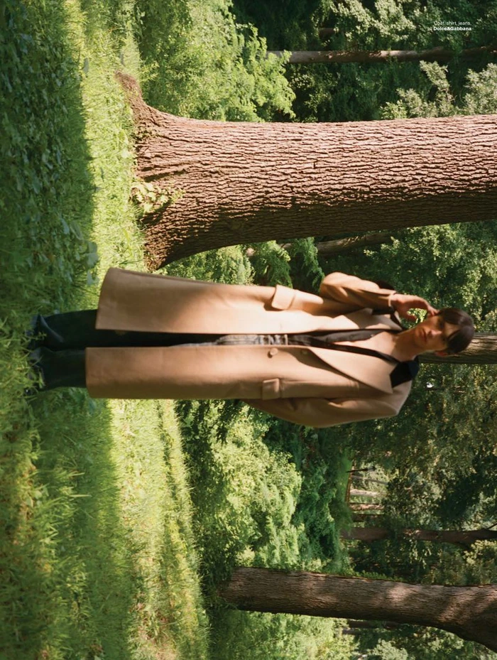 Model wearing a long camel coat posing outdoors near a large tree trunk in a park setting.