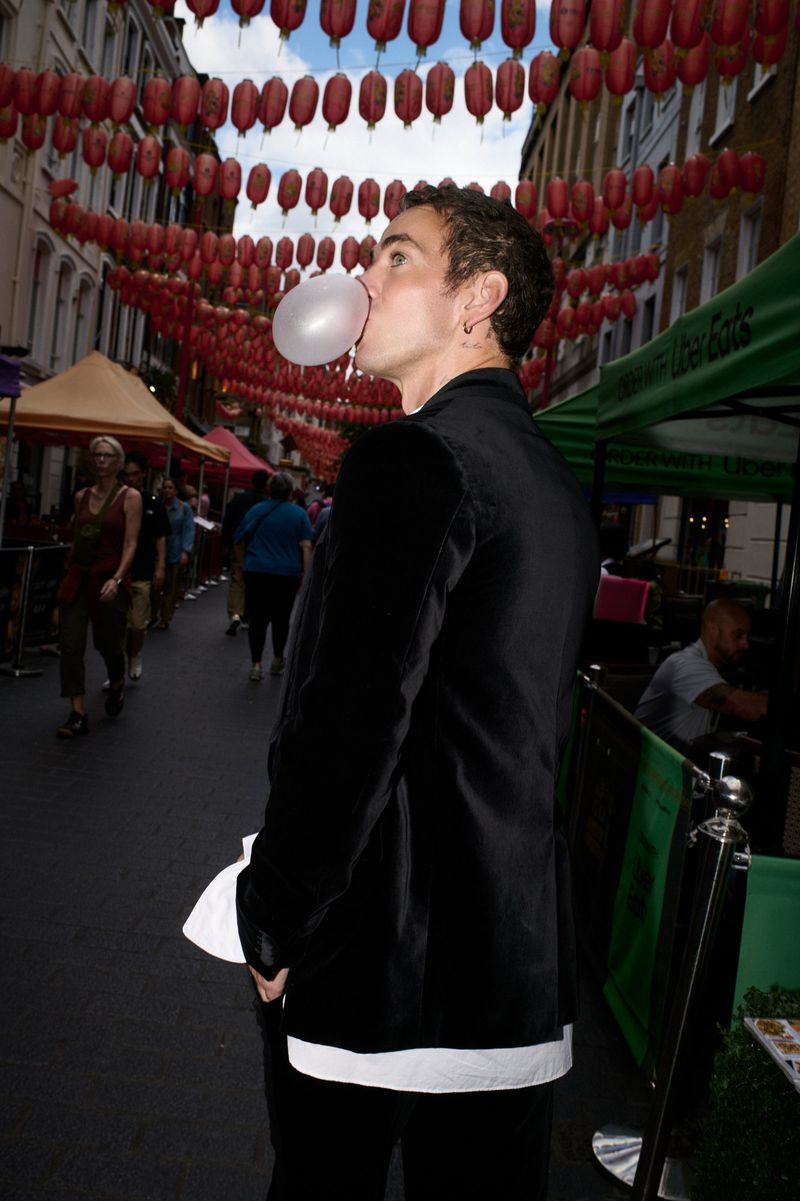 Man in a black velvet blazer blowing a bubble with gum on a bustling street lined with red lanterns.