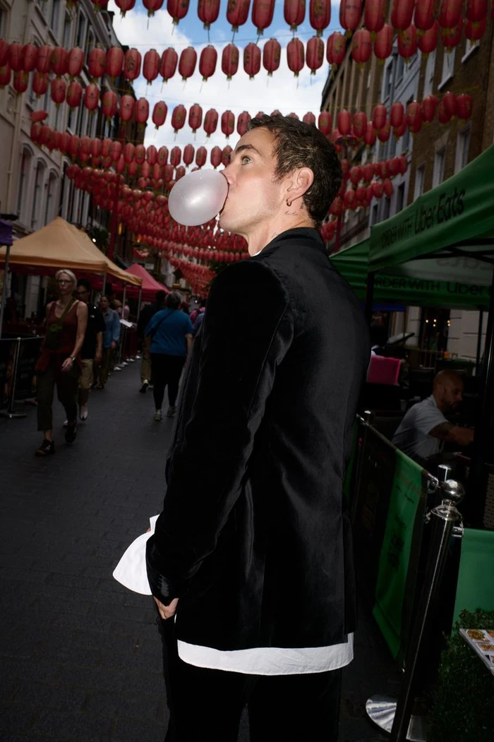 Man in a black velvet blazer blowing a bubble with gum on a bustling street lined with red lanterns.