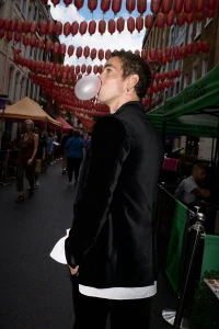 Man in a black velvet blazer blowing a bubble with gum on a bustling street lined with red lanterns.