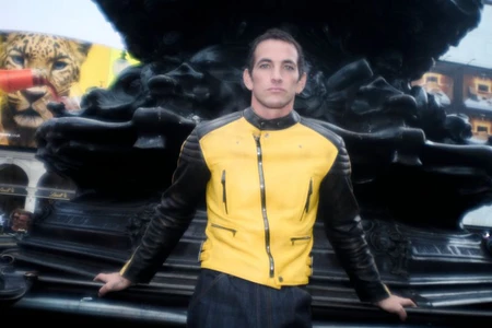 Alex Fine posing in front of an ornate dark sculpture wearing a yellow and black leather jacket for Numero Netherlands.