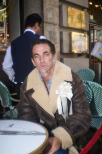 Man wearing brown shearling coat seated at a café table with a white flower lapel pin
