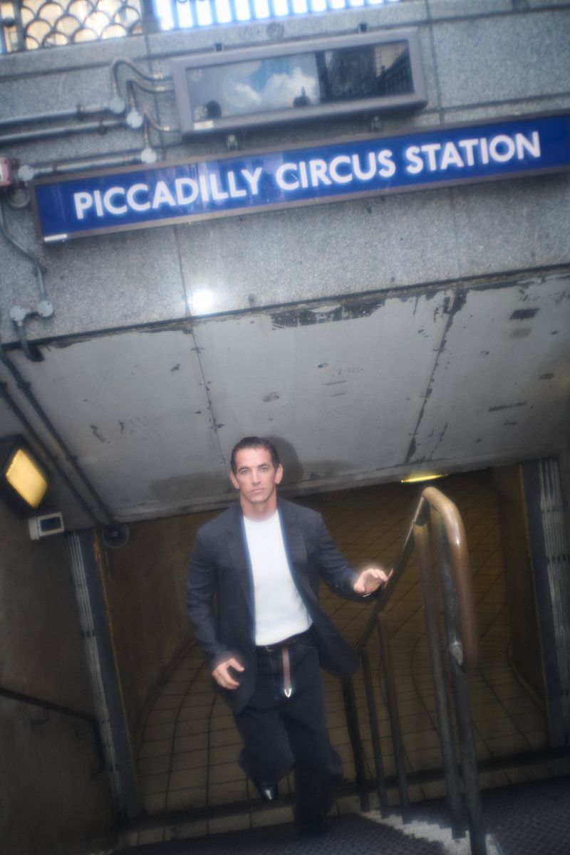 Man wearing a dark jacket and white t-shirt ascending stairs beneath a Piccadilly Circus Station sign