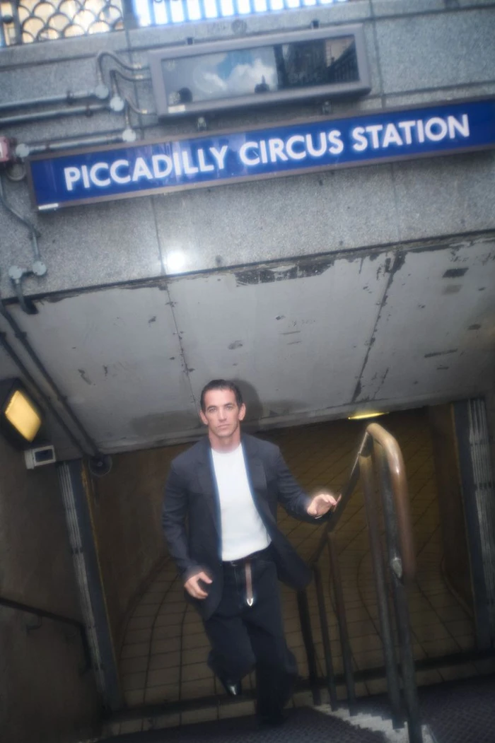 Man wearing a dark jacket and white t-shirt ascending stairs beneath a Piccadilly Circus Station sign