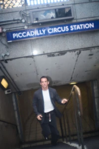 Man wearing a dark jacket and white t-shirt ascending stairs beneath a Piccadilly Circus Station sign