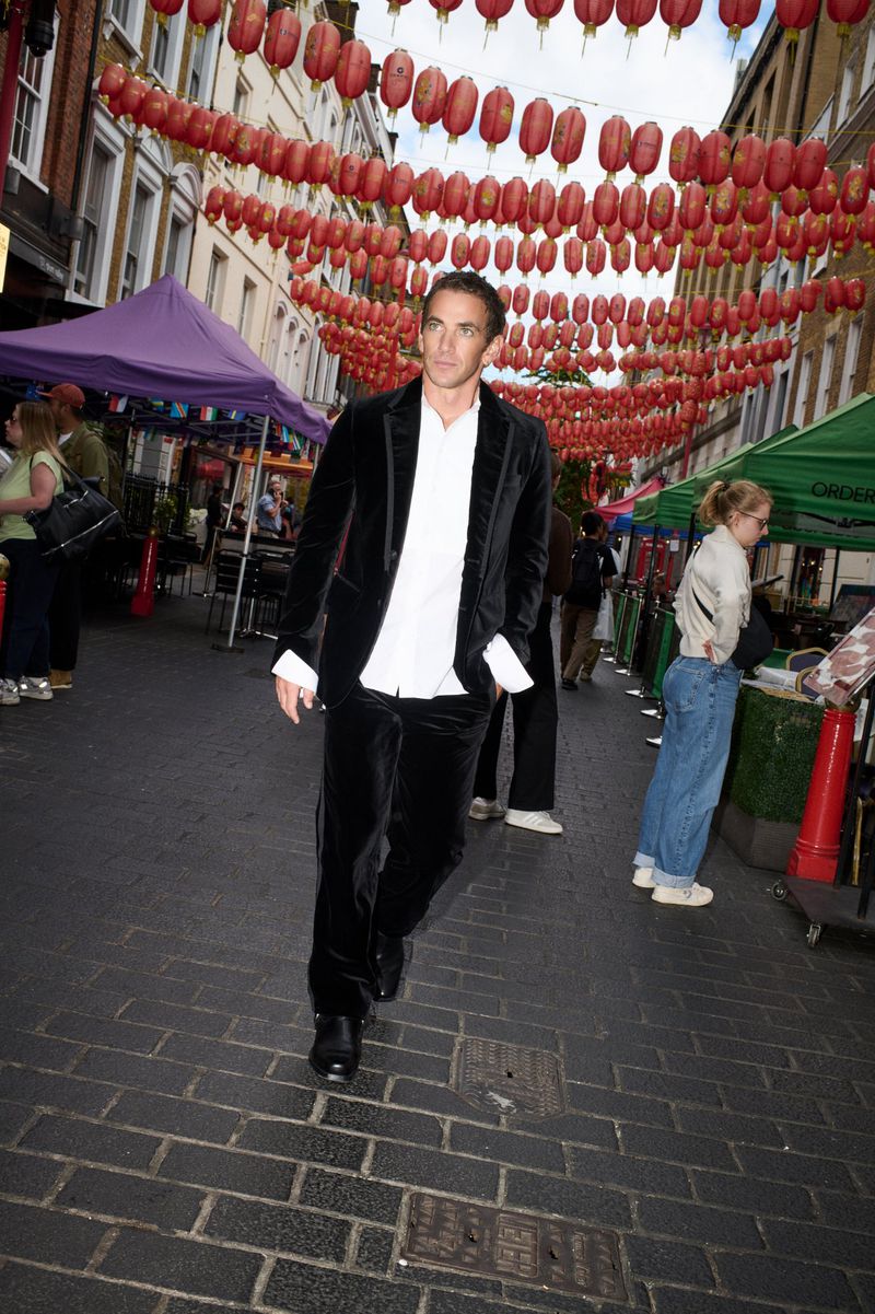 Man walking on a street decorated with red lanterns, wearing a black velvet suit and unbuttoned white shirt.