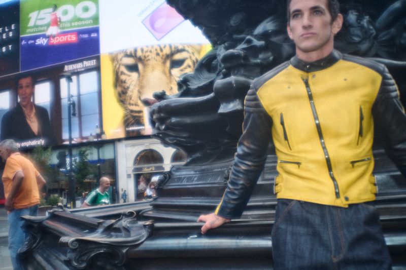 Man in a yellow and black leather jacket posing near an outdoor public monument with large billboards in background