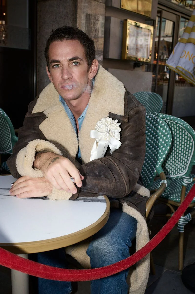 Man wearing a shearling-lined brown coat and blue shirt seated at an outdoor café table exhaling a wisp of smoke.