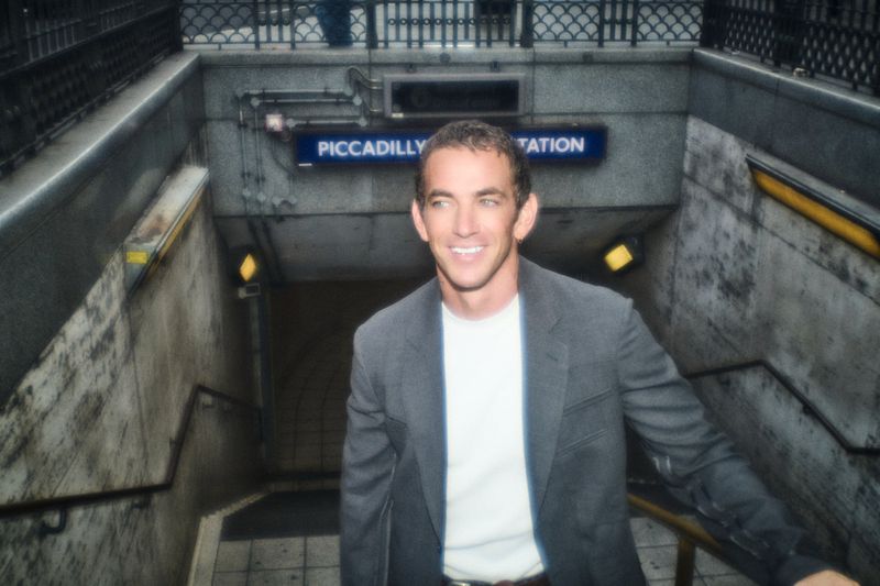 Alex Fine wearing a grey blazer and white t-shirt ascending the steps at Piccadilly Circus station