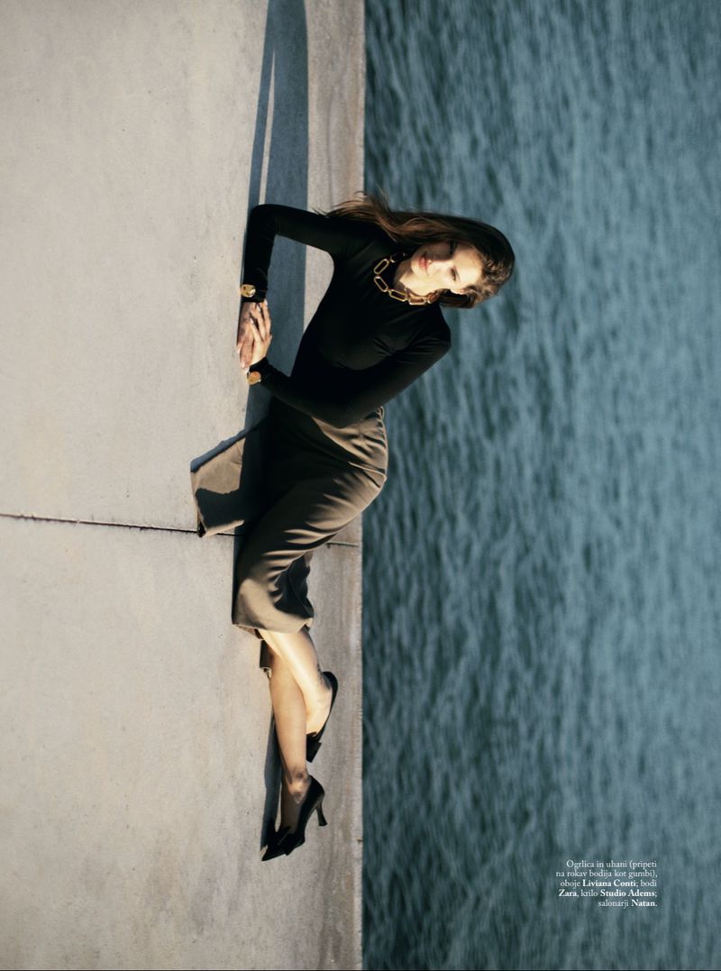 Model Faretta wearing black top and midi skirt posing by the water for Elle Slovenia, photographed by Suzana Holtgrave.