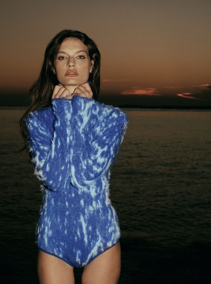 Model Faretta wearing a patterned blue knit top standing by the water at dusk for Elle Slovenia