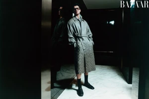 Model standing in a hallway wearing a grey trench coat and dark socks with a reflection in a glass wall