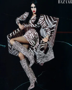 Model wearing zebra-print clothing and boots seated in matching armchair against dark background