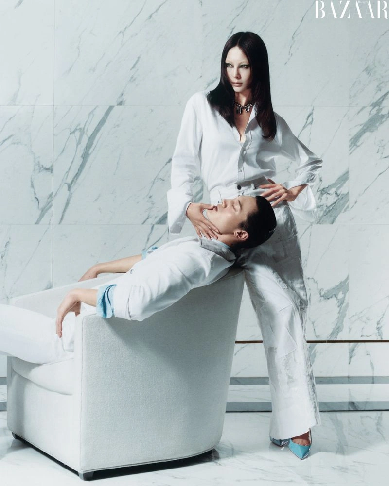 Two models in white attire with one standing and resting her hand on the seated person's chin against a marble wall