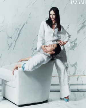 Two models in white attire with one standing and resting her hand on the seated person's chin against a marble wall