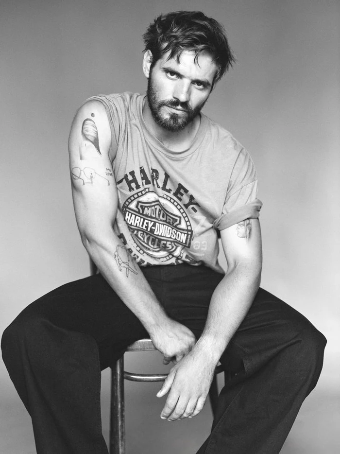 Portrait of Tyler Conti seated on a stool wearing a printed t-shirt and dark trousers against a seamless neutral backdrop.