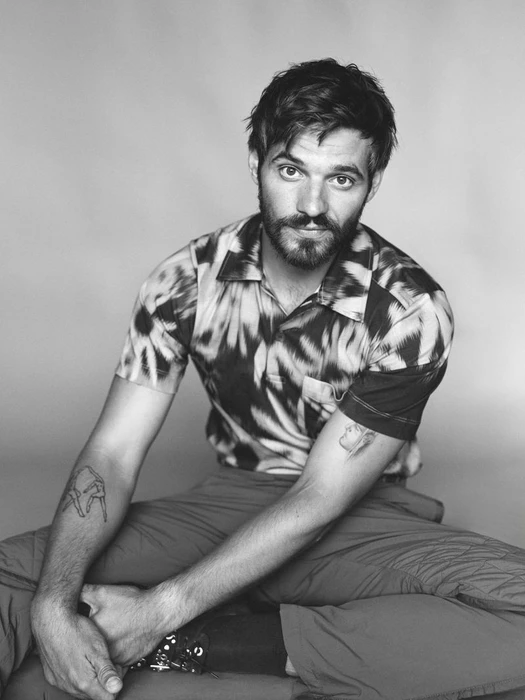 Portrait of Tyler Conti seated against a plain light background wearing a patterned polo shirt and tattoos on his forearms.