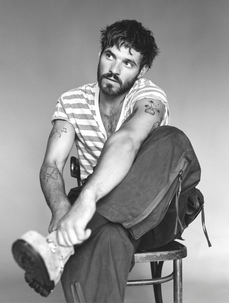 Monochrome portrait of Tyler Conti seated on a chair wearing a striped t-shirt and dark trousers, looking upwards.