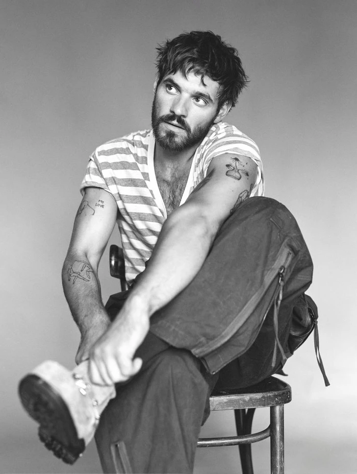Monochrome portrait of Tyler Conti seated on a chair wearing a striped t-shirt and dark trousers, looking upwards.
