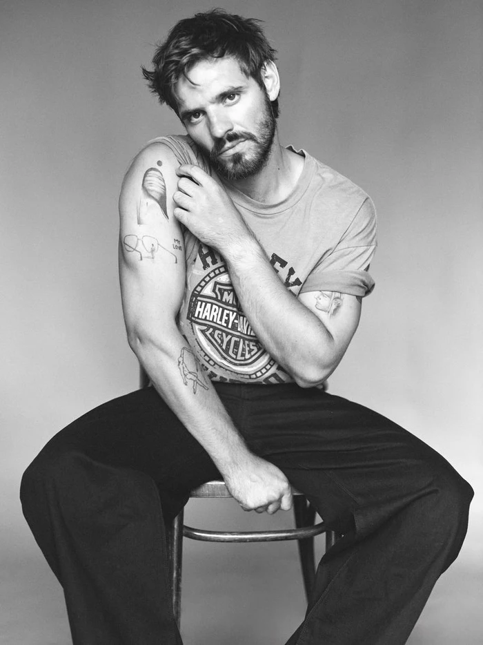 Man with tattoos and a beard seated on a stool wearing a t-shirt against a grey seamless background