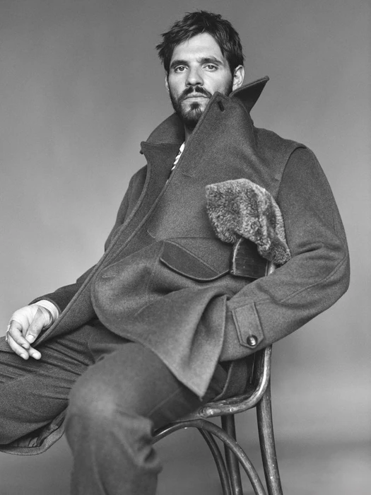 Man with beard wearing a heavy wool coat seated on a chair against a neutral background