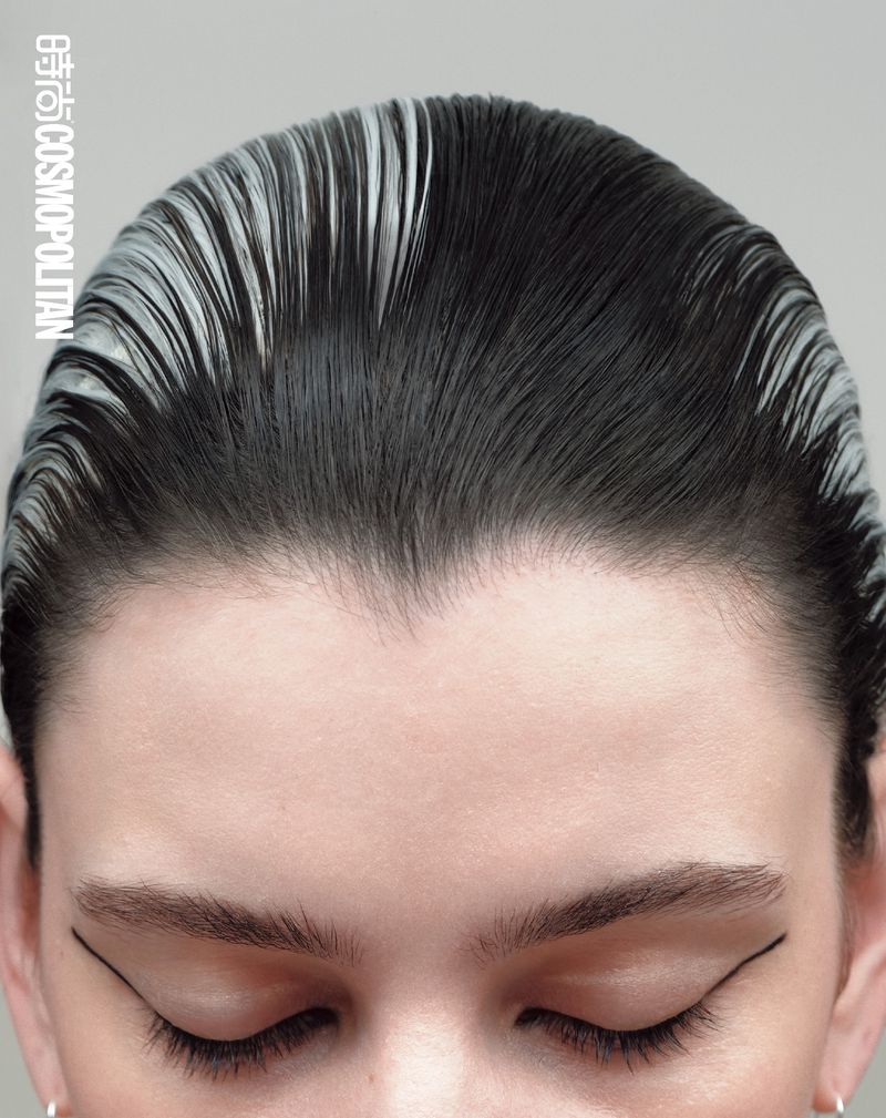 Close-up of a light-skinned adult female's face with slicked back dark hair with silver roots and strong graphic black eyeliner, eyes closed.