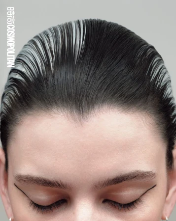 Close-up of a light-skinned adult female's face with slicked back dark hair with silver roots and strong graphic black eyeliner, eyes closed.
