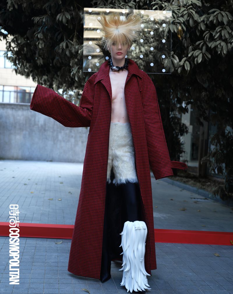 Fashion-forward woman in a red coat with avant-garde hair and unique boots posing outdoors, blending high fashion with urban style.