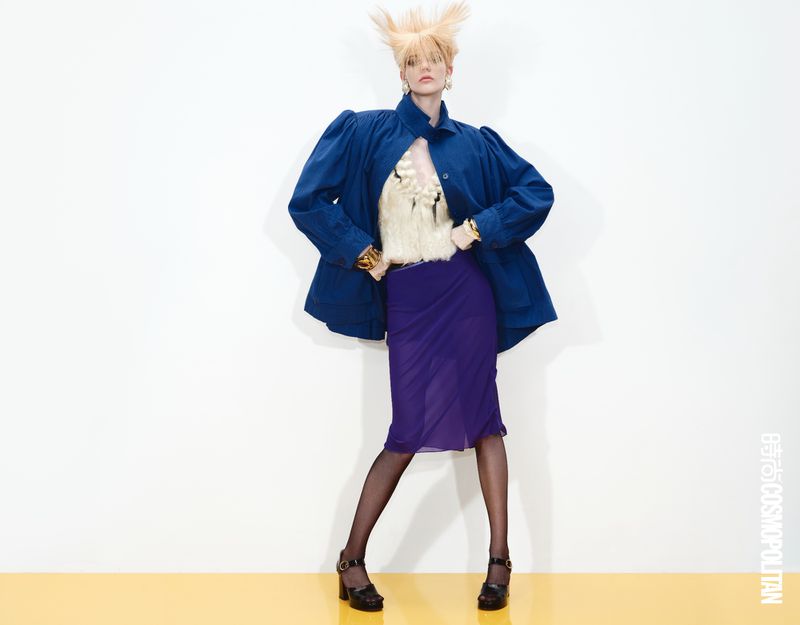 A woman with blonde hair poses in a blue jacket, purple skirt, and black shoes in a studio with a yellow floor and white background.