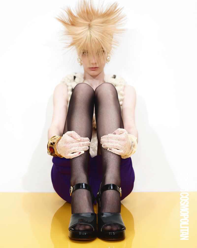 A woman with wild blonde hair and platform heels sits on a yellow surface in a studio against a white background. She wears black tights.