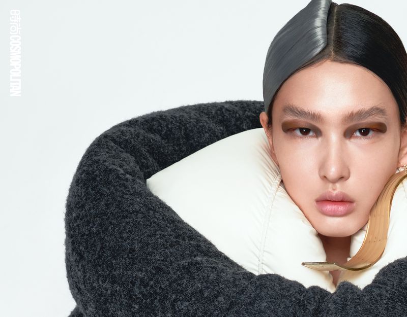 Close-up studio portrait of a model with gray and black parted hair, bold eye makeup, white and gray puffy coat, and large gold earring.