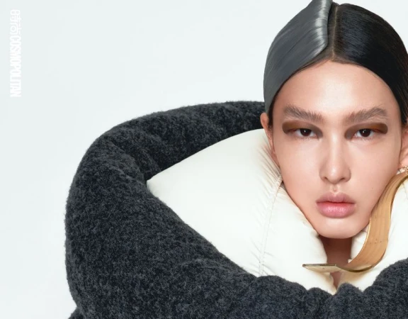 Close-up studio portrait of a model with gray and black parted hair, bold eye makeup, white and gray puffy coat, and large gold earring.