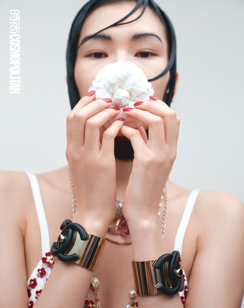 Elegant Asian woman with bold makeup poses indoors with white flower and jewelry, wearing decorative bracelets, fashion editorial shot.