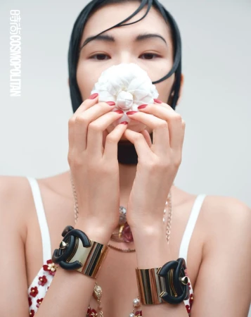 Elegant Asian woman with bold makeup poses indoors with white flower and jewelry, wearing decorative bracelets, fashion editorial shot.