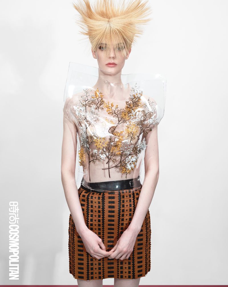 Fashion photo of a model with blonde spiky hair wearing a transparent top with tree branch designs and a brown skirt on a white background.