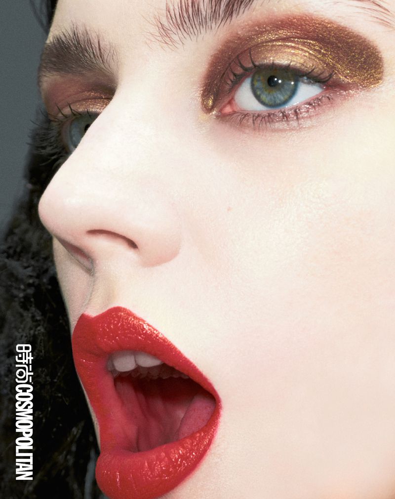 Close-up shot of a woman's face featuring bold makeup with red lipstick, golden eye shadow, and manicured eyebrows, set against a soft backdrop.
