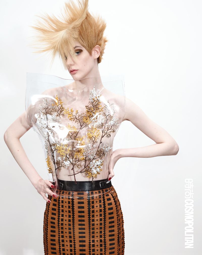 Fashionable young woman with blonde updo hair poses in clear, embellished floral top and brown patterned skirt against a white background.