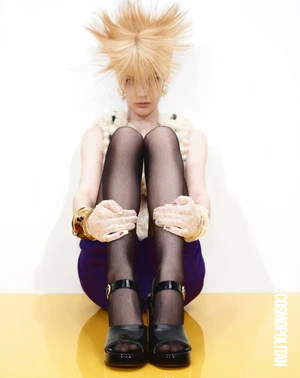 Model with spiky blonde hair sitting on a yellow surface wearing black tights and platform shoes