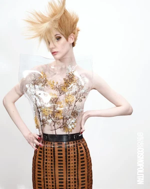 Model with spiky blonde hair wearing a clear plastic bodice enclosing branches and a patterned brown skirt against a white background.